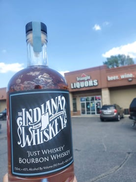 A bottle of whiskey that’s in front of a liquor shop named “Triangle Liquors” beside a sign that reads, “beer. wine.”.