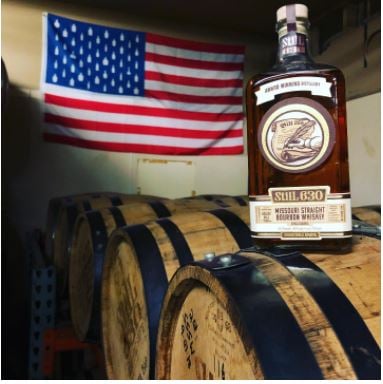 A bottle of  whiskey placed on top of whiskey barrels with the American flag hanging on the wall behind it.