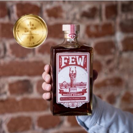 A bottle of FEW Spirit’s American Straight Whiskey held by one hand in front of a brick wall background.