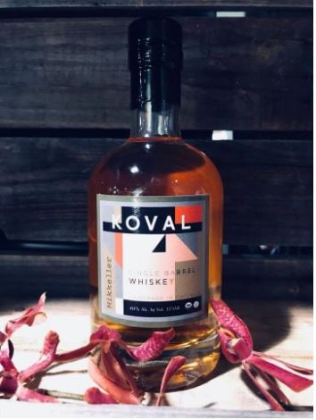 A bottle of KOVAL Mikkeller Single Barrel Whiskey on top of a wooden bench with flowers in the foreground.