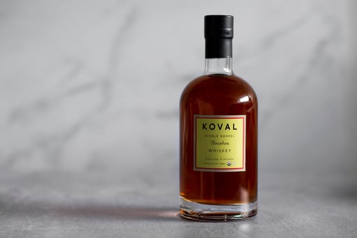 A bottle of KOVAL Bourbon Whiskey placed on a white marble floor with a blurred marble background.