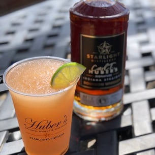 A bottle of Bottled-In-Bond Carl T. Bourbon Whiskey on a table beside a full glass of an iced drink with lime.