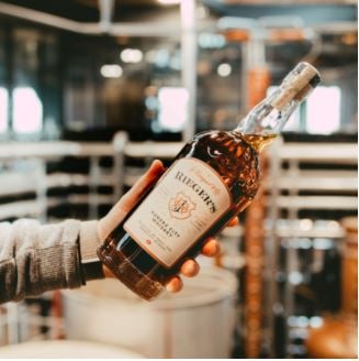 Someone standing out of frame holds a bottle of Rieger’s Kansas City Whiskey inside a distillery.