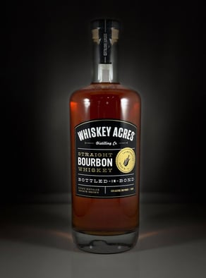 A bottle of Whiskey Acres’ Bottled in Bond Bourbon on a black table with a black background.