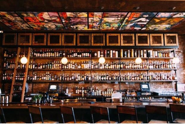 Well-lit bar, a line of chairs in front of the counter with wooden shelves behind the counter filled with spirits.