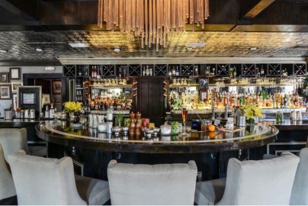 Brightly lit bar with white chairs, shelves are filled with spirits and brightly lit chandelier.  