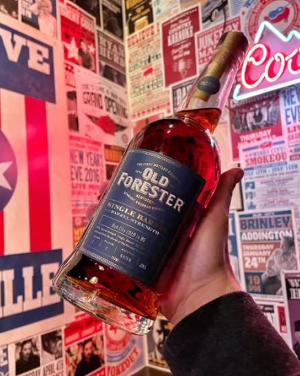 Image of a bottle of old forester single barrel bourbon with a backdrop of a variety of posters stuck on the wall
