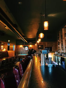 Sideways view of whiskey bar with lights turned on and hanging above the counter with maroon chairs in front.