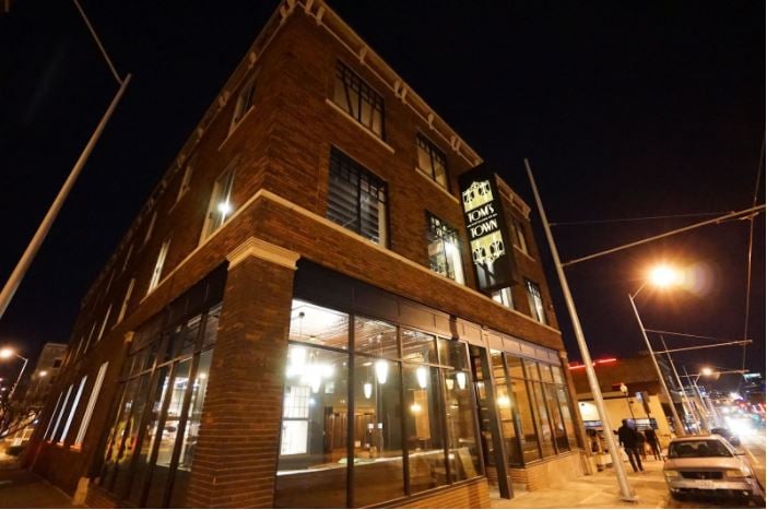 An image of Tom’s Town Distilling Co.’s facade; the lights are on, and people are walking on the streets at night