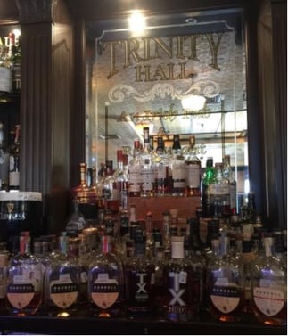 Trinity Hall Irish Pub’s spirits on display. Behind the bottles is signage that reads, “Trinity Hall, An Irish Pub.